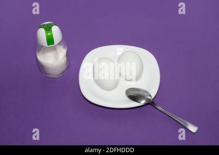 Deux œufs blancs sur une assiette. Un plat de deux œufs avec une cuillère et du sel sur fond violet. Œufs durs pour le petit déjeuner. Banque D'Images