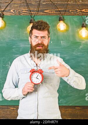 Enseignant dans les lunettes est titulaire d'un réveil. Régime de l'annexe et concept. Hipster barbu est titulaire d'réveil, tableau sur toile de fond, l'espace de copie. Homme avec barbe et moustache sur surpris face au réveil de pointage Banque D'Images