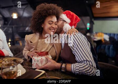 beardy caucasien gars embrassant dans la joue afro-américaine femelle, chuchotant à l'oreille, lui donnant un cadeau de noël, portant chapeau de santa Banque D'Images