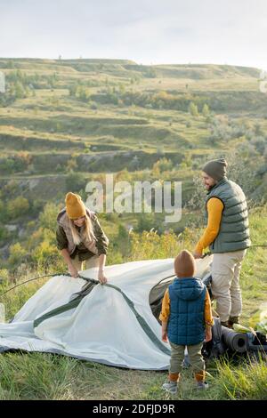 Jeune couple et leur petit fils debout au-dessus de montagne tout en mettant la tente sur le sol devant caméra pendant le week-end Banque D'Images