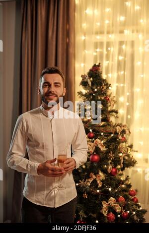 Joyeux jeune barbu homme en chemise blanche tenant la flûte de champagne tout en se tenant contre un arbre de Noël décoré pendant la fête de la maison Banque D'Images