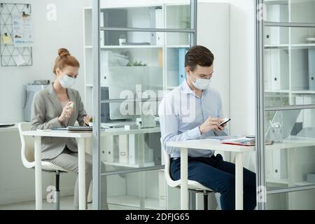 Jeune homme d'affaires dans le masque de protection et de tenue de forme en utilisant le smartphone pendant sa collègue vaporisant de l'assainisseur sur les mains derrière lui Banque D'Images