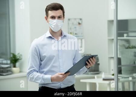 Jeune homme d'affaires en masque de protection et de tenue de forme vous regardant lors de l'utilisation d'un document financier dans le presse-papiers, devant appareil photo Banque D'Images