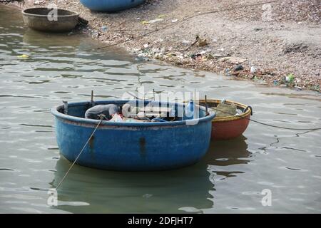 Le pêcheur vietnamien vérifie les fournitures dans son bateau en fibre de verre tandis que les coracles à armure de panier traditionnel bob à proximité. Banque D'Images