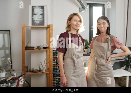 Deux jeunes artisans interculturels à succès en vêtements de travail à proximité une autre face à la caméra dans un grand atelier ou studio Banque D'Images