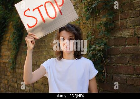 Une jeune femme protestataire en chemise blanche et un Jean tient un écriteau de l'enseigne de protestation avec le slogan « Stop » pour une manifestation publique sur fond de mur. Banque D'Images