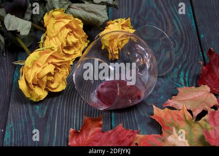 Un bouquet de roses jaunes séchées et de feuilles d'automne séchées. Un verre de vin aigre renversé. Sur les planches de pin. Banque D'Images