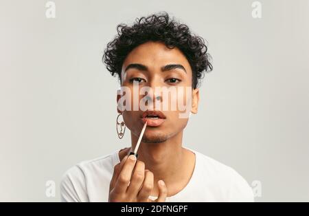 Jeune homme appliquant le brillant à lèvres sur ses lèvres et regardant dans l'appareil photo comme un miroir. Homme gay faisant le maquillage sur fond blanc. Banque D'Images