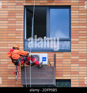 Un ouvrier masculin en uniforme rouge installe un climatiseur dans une niche sous la fenêtre - Moscou, Russie, 18 juin 2020 Banque D'Images