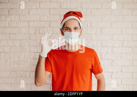 Jeune homme messagerie dans le masque protecteur, gants médicaux, chapeau de père noël montrer signe ok et sourire. Heureux accouchement homme dans médical masque gestuel OK symb Banque D'Images