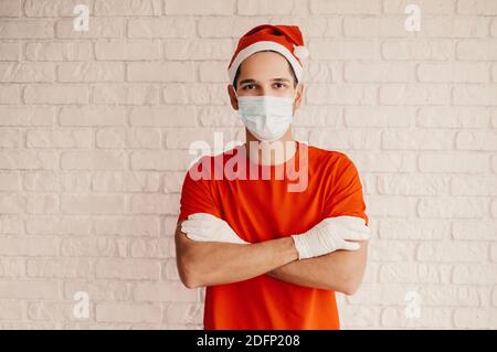 Jeune employé de service de livraison professionnelle à santa chapeau, masque médical, gants de protection garder les bras croisés, sourire. Joyeux homme confiant messager Banque D'Images