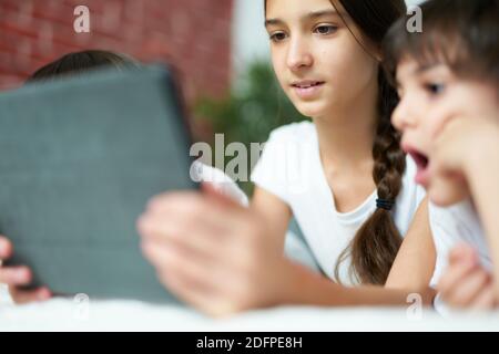 Portrait en gros plan de la jolie fille latine souriant en utilisant une tablette numérique. Sœur adolescente qui divertit ses deux petits frères, regardant des dessins animés à la maison. Bonne enfance, concept de technologie Banque D'Images