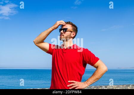 Jeune homme expressif dynamique ambitieux réussi barbu en t-shirt rouge lunettes de soleil en été bleu ciel océan fond. Concept de liberté de style de vie Banque D'Images