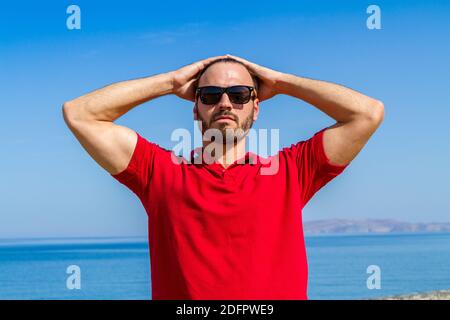 Jeune homme expressif dynamique ambitieux réussi barbu en t-shirt rouge lunettes de soleil en fond bleu ciel d'été. Banque D'Images