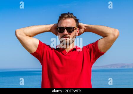 Jeune homme expressif dynamique ambitieux réussi barbu en t-shirt rouge lunettes de soleil en été bleu ciel océan fond. Concept de liberté de style de vie Banque D'Images