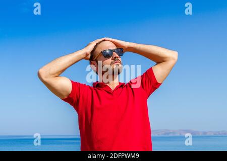 Jeune homme expressif dynamique ambitieux réussi barbu en t-shirt rouge lunettes de soleil en été bleu ciel océan fond. Concept de liberté de style de vie Banque D'Images