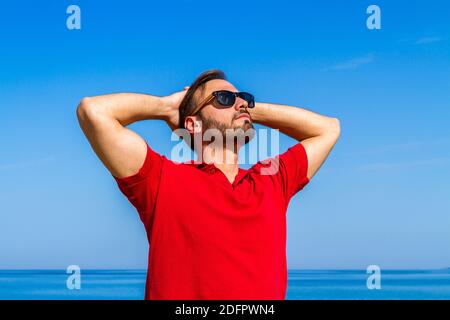 Jeune homme expressif dynamique ambitieux réussi barbu en t-shirt rouge lunettes de soleil en été bleu ciel océan fond. Concept de liberté de style de vie Banque D'Images