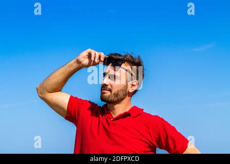 Jeune homme expressif dynamique ambitieux réussi barbu en t-shirt rouge lunettes de soleil en été bleu ciel océan fond. Concept de liberté de style de vie Banque D'Images