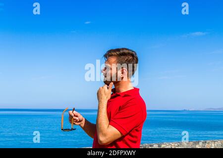 Jeune homme expressif dynamique ambitieux réussi barbu en t-shirt rouge lunettes de soleil en été bleu ciel océan fond. Concept de liberté de style de vie Banque D'Images