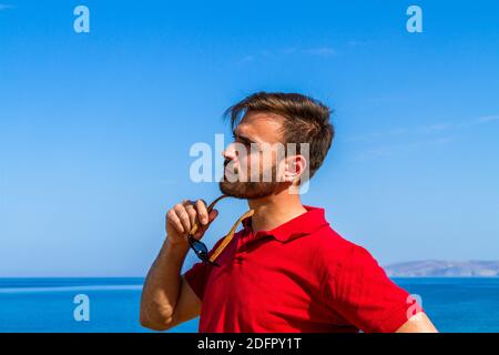 Jeune homme expressif dynamique ambitieux réussi barbu en t-shirt rouge lunettes de soleil en été bleu ciel océan fond. Concept de liberté de style de vie Banque D'Images