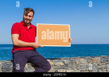 Jeune barbu beau homme souriant habillé en bleu rouge décontracté avec un geste invitant, tenant le bois encadré blanc corkboard dans bleu ciel bac océan Banque D'Images