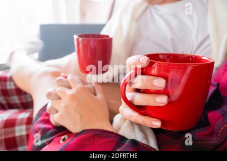 Tasse rouge à la main. Vue latérale courte d'un homme et d'une femme tenant les mains assis à la maison sur un canapé en pyjama écossais Banque D'Images