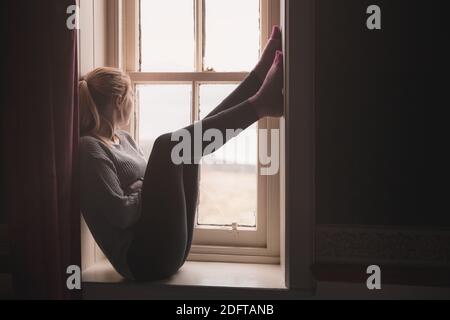 Une jeune femme coincée à l'intérieur s'assoit sur un seuil de fenêtre, regardant à l'extérieur et aspirant à être libre. Banque D'Images