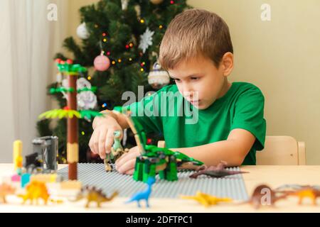 SAINT-PÉTERSBOURG, RUSSIE - 29 NOVEMBRE 2020 Boy construit un monde jurassique à partir de blocs de plastique et joue avec les dinosaures de jouets. Arbre de Noël sur fond. Restez à la maison pour les vacances de Noël concept Banque D'Images