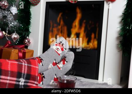 Gros plan des jambes des filles près de la cheminée avec une flamme et une tasse de thé sur le sol à la maison. Les jambes des femmes sont recouvertes d'une couverture et portent des chaussettes décoratives de Noël Banque D'Images