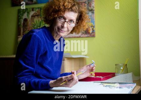Bonne femme d'artiste mûre peint sur toile à la maison studio. Exprimez votre créativité. Femme âgée positive tenant la brosse et dessin sur la toile pendant Banque D'Images