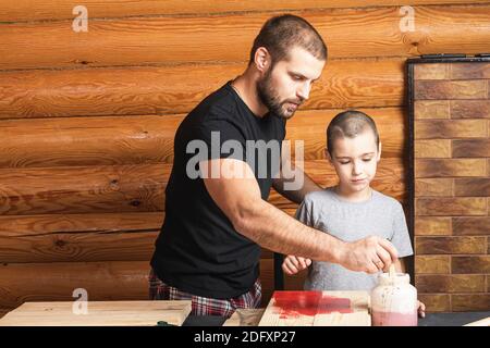 Le père et le fils forment ensemble une maison d'oiseaux en bois dans l'atelier. Un père joyeux avec un petit garçon peint un panneau de bois avec une brosse en rouge sur un tabl Banque D'Images