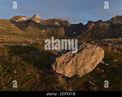 Grand rocher couché sur le sol sur un pré crié avec des buissons colorés, de l'herbe et de la mousse jetant l'ombre dans la lumière du soir avec des montagnes en arrière-plan. Banque D'Images