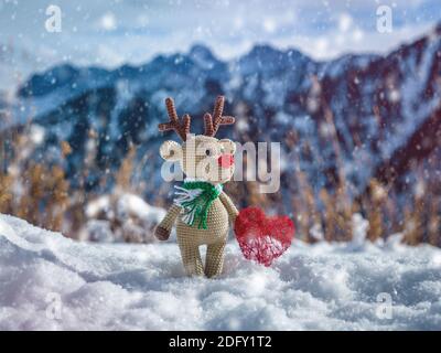Un triste cerf jouet avec un coeur dans ses pattes est en hiver dans les montagnes en chute de neige. Jouet de Noël fait main Banque D'Images