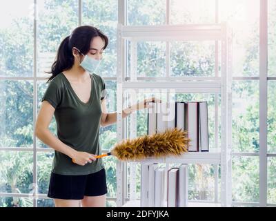Jeune belle femme asiatique, femme au foyer en tissu décontracté portant masque de protection livres de nettoyage sur la bibliothèque avec plumeau dans le salon sur Banque D'Images