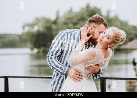 Les beaux jeunes mariés posent sur la jetée. Le marié épouse doucement et embrasse le cou de la mariée sur le quai. Banque D'Images