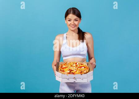 Portrait d'une femme sportive positive tenant une boîte en carton avec une pizza grasse, appréciant la nourriture de la malbouffe après l'entraînement dans la salle de gym, manque de volonté. Salle de studio intérieure Banque D'Images