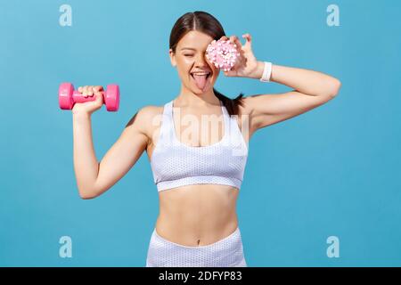 Drôle de sport positif femme dans les collants et le haut avoir l'amusement, la fermeture de l'œil avec le donut et montrant la langue, tenant la haltère dans la main, brûlant des calories supplémentaires. Banque D'Images