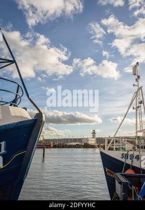 Un phare et un quai sont aperçus entre les arcs de deux chalutiers qui font face l'un à l'autre. Un ciel bleu est au-dessus avec des nuages. Banque D'Images