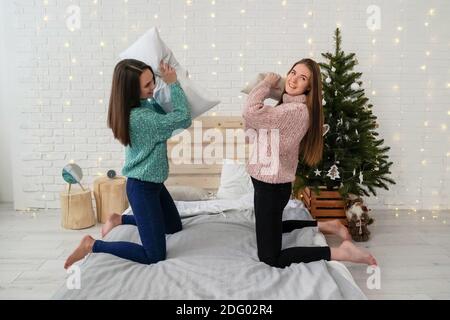 Deux jeunes femmes dans des chandails ayant du plaisir, combat d'oreiller sur le lit. Humeur des fêtes. Banque D'Images