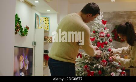 Zoom sur la photo d'arbre de noël assemblé par un jeune couple dans le salon. Décoration de beaux arbres de Noël avec des décorations de boule en verre. Femme et mari dans des vêtements assortis aidant à la maison ornée avec guirlande lumineuse Banque D'Images