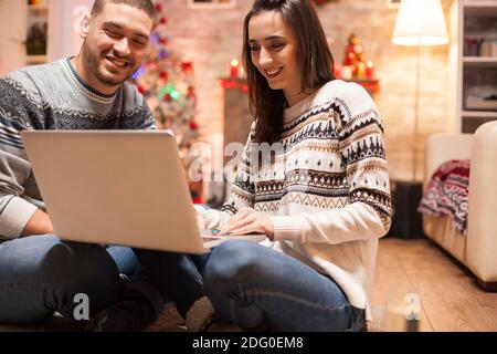 Couple heureux s'amuser sur Internet en utilisant un ordinateur portable le jour de noël, devant une cheminée chaude. Banque D'Images