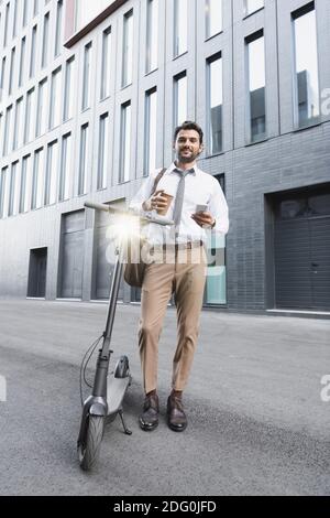 pleine longueur d'homme d'affaires gai en costume tenant une tasse en papier et en utilisant un smartphone près d'un e-scooter Banque D'Images
