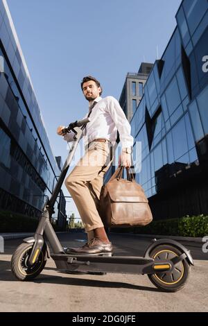 vue à angle bas de l'homme d'affaires barbu dans la tenue habillée sac en cuir et debout près du scooter électrique Banque D'Images