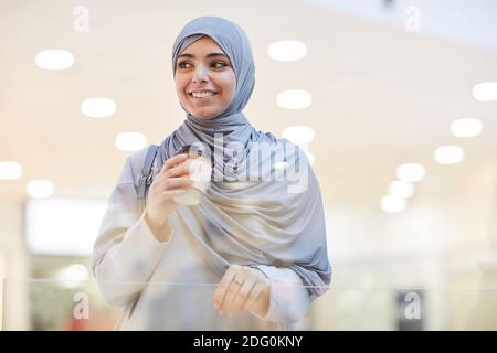 Portrait à la taille haute d'une jeune femme du Moyen-Orient portant un foulard et tenant une tasse de café tout en appréciant les magasins en ville, espace copie Banque D'Images