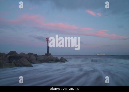 Des nuages roses rayent dans le ciel au coucher du soleil et des vagues s'écrasées sur la rive autour de la base d'une groyne pendant qu'un bateau de croisière se trouve à l'horizon. Banque D'Images