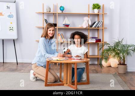 Pleine longueur de psychologue multiculturel heureux et fille regardant appareil photo assis près d'une table basse dans le bureau Banque D'Images