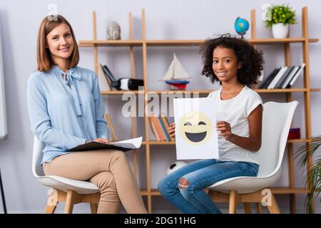 Une fille afro-américaine souriante montrant du papier avec une expression heureuse pendant assis près d'un psychologue sur fond flou Banque D'Images