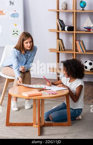 Pleine longueur de psychologue regardant sourire fille afro-américaine dessin tout en étant assis près d'une table basse dans le bureau Banque D'Images