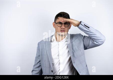 Jeune homme d'affaires portant une chemise décontractée sur fond blanc très heureux et souriant regardant loin avec la main sur la tête. Concept de recherche. Banque D'Images