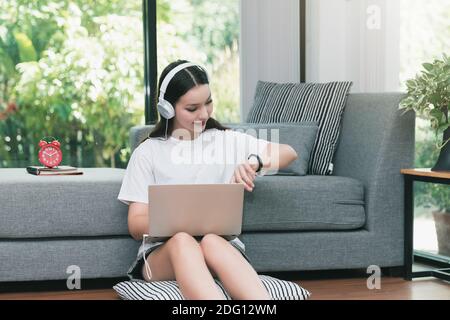 Jeune femme asiatique avec casque regarde la montre intelligente tout en étant assise sur le sol dans le salon. Cours en ligne avec un ordinateur portable à la maison. Banque D'Images